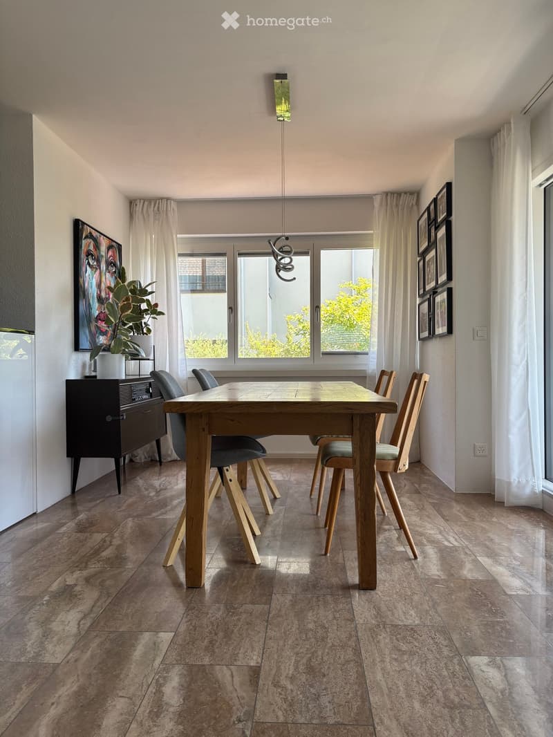 Modern kitchen with a wooden dining table, four chairs, a dark wooden cabinet, a potted plant, and several picture frames on the walls. Large windows with white curtains allow natural light to fill the space.