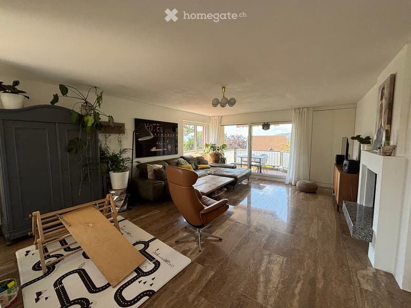 Bright living room with large windows and a sliding glass door leading to a balcony. Features a grey sectional sofa, a wooden coffee table, a leather armchair, a dark cabinet, a fireplace, and a TV screen. The room includes a patterned rug with a road des