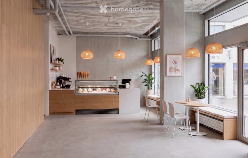 Modern, minimalist interior with pendant lights, white tables, chairs, a bench, and a counter with a coffee machine and display case.