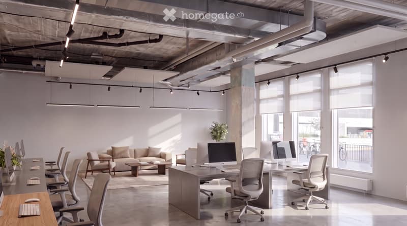 Modern office space with exposed ceiling beams, large windows, desks, chairs, a sofa, coffee table, and computer monitors.