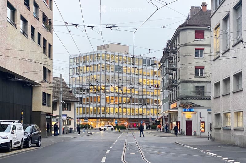 Urban street scene with modern glass building, traditional buildings, pedestrians, and vehicles.