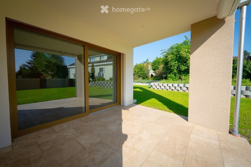 Spacious balcony with tiled flooring, large sliding glass doors, and a view of a well-maintained garden with a stone wall and trees.