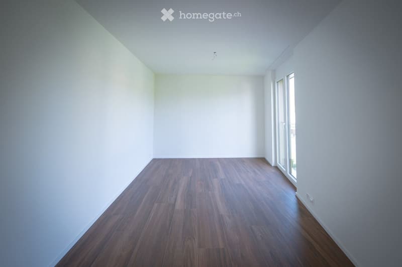 Empty room with wooden flooring and white walls, featuring large windows on one side.