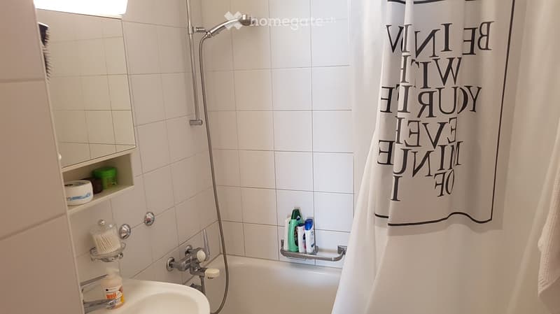 Modern bathroom with white tiles, a bathtub, showerhead, mirror, and shelves holding toiletries.