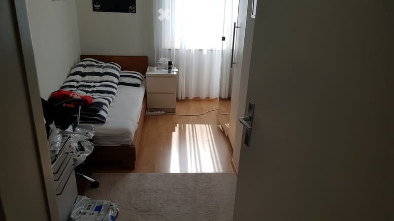 A small, furnished bedroom with a single bed, nightstand, and window with curtains. The room has wooden flooring and a light-colored rug.