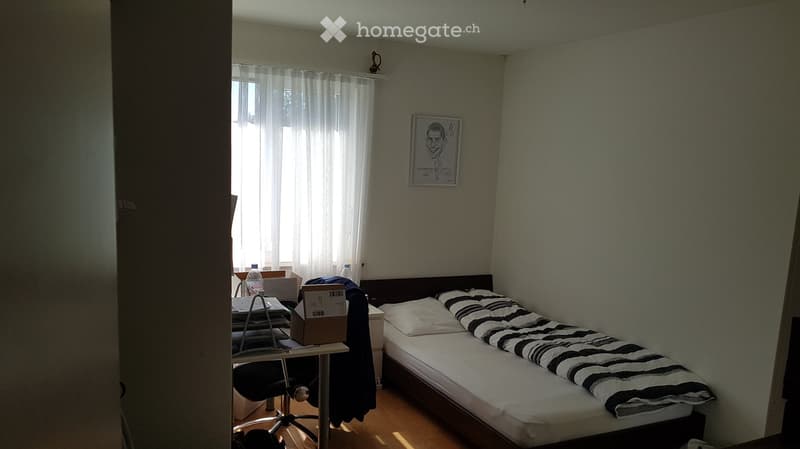 Small bedroom with a single bed, a chair, a small table, and a picture frame on the wall. The bed has a striped blanket and white pillows. There is a window with white curtains allowing natural light to enter.