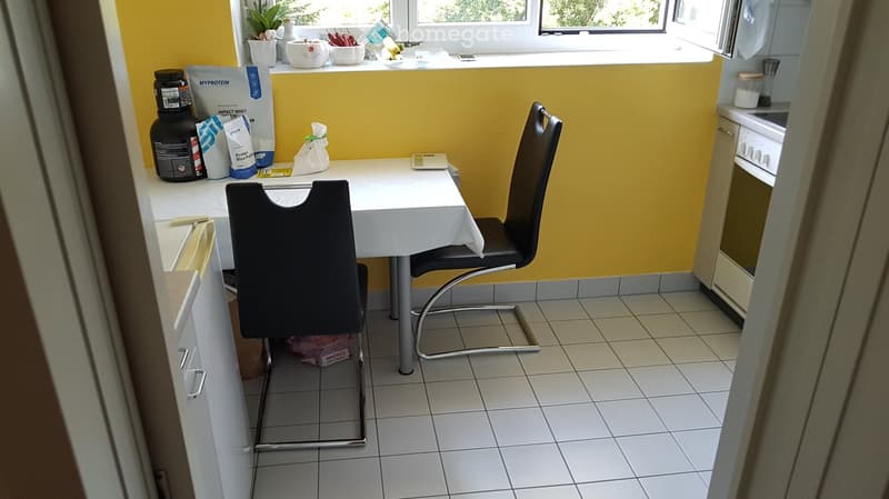 Bright kitchen with yellow walls, white tiled floor, and a small dining area with a table and two chairs. Visible items include a blender, a cup, and some bags of food on the counter.