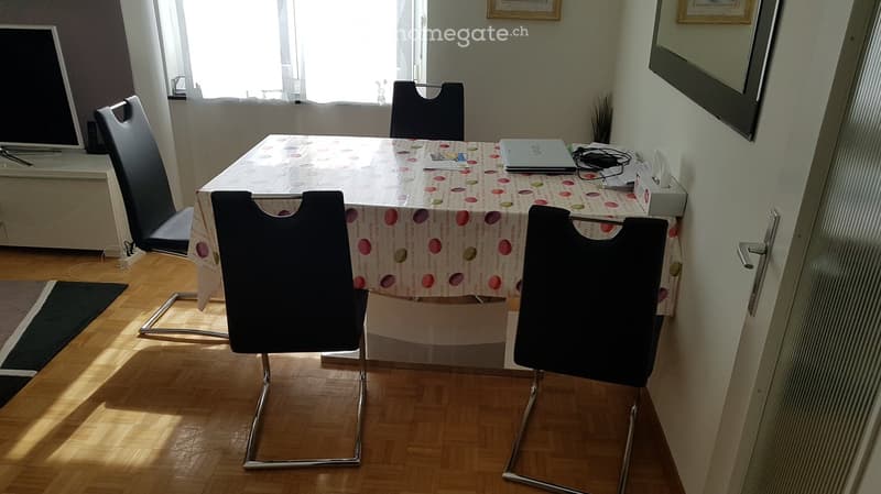 Bright room with wooden flooring, a table covered with a colorful tablecloth, four black chairs, a laptop, and a TV screen visible in the background.