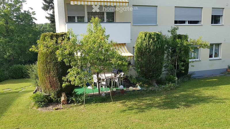 Well-maintained garden with green lawn, patio furniture, small trees, and shrubs surrounding a two-story house with balconies and striped awnings.