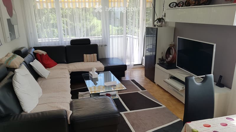 Modern living room with a black leather sofa, glass coffee table, TV stand, and decorative items on shelves.