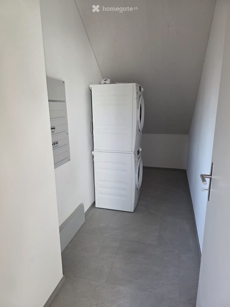 A small, narrow room with a washing machine and dryer stacked vertically, white walls, and a grey tiled floor.