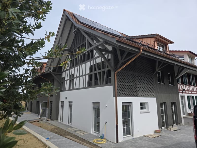 Modern two-story house with grey and white exterior, large windows, and a balcony on the upper level. Solar panels are installed on the roof.