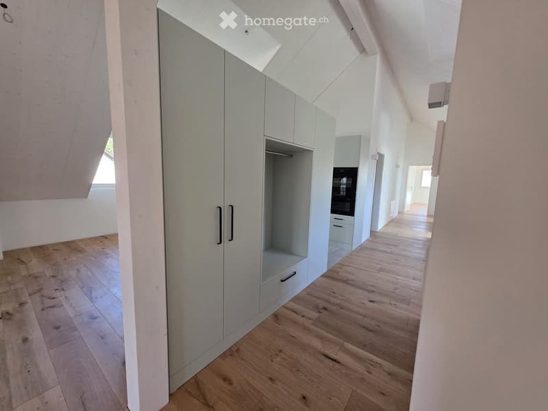 Bright, modern hallway with wooden flooring, white walls, and built-in light grey storage units with glass doors and black handles. The hallway has a slanted ceiling with skylight windows, leading to another room with visible kitchen appliances.