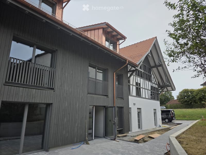 Modern two-story house with dark wooden siding, large windows, and a balcony on the upper level. The house features a red-tiled roof and a contemporary design with a mix of dark and light elements.