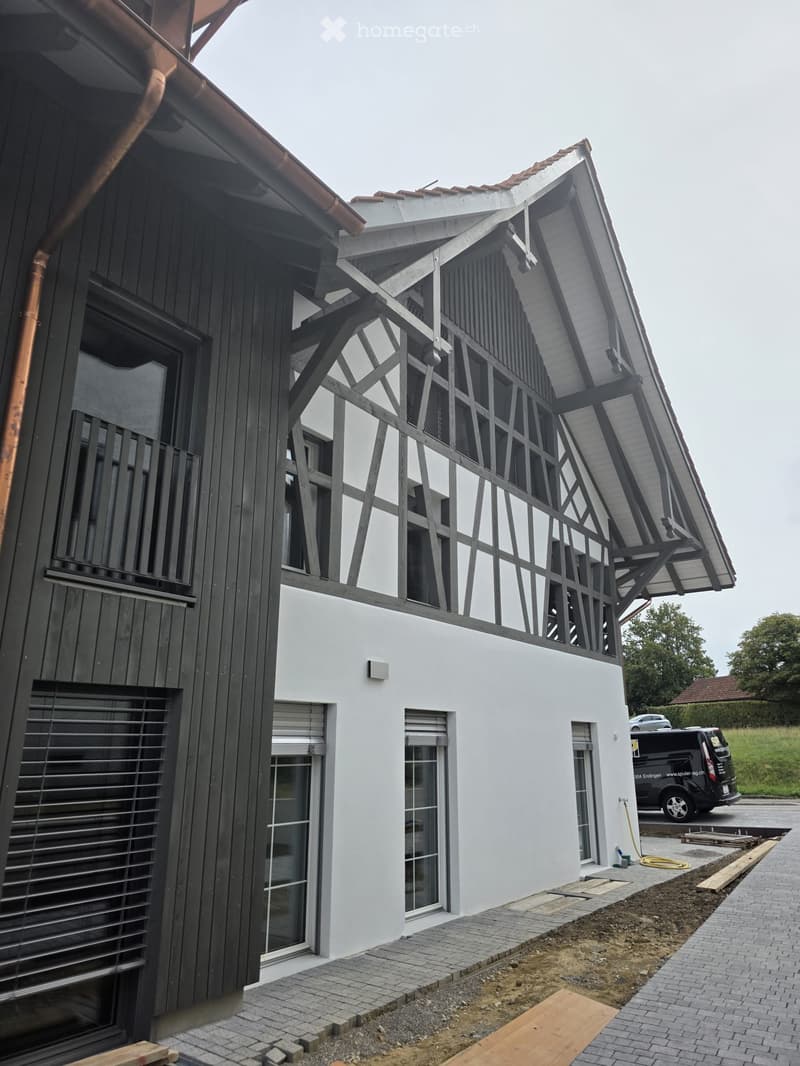 Modern two-story house with white and black exterior, large windows, and a distinctive roof design. The ground floor features a covered entrance area with a paved walkway.
