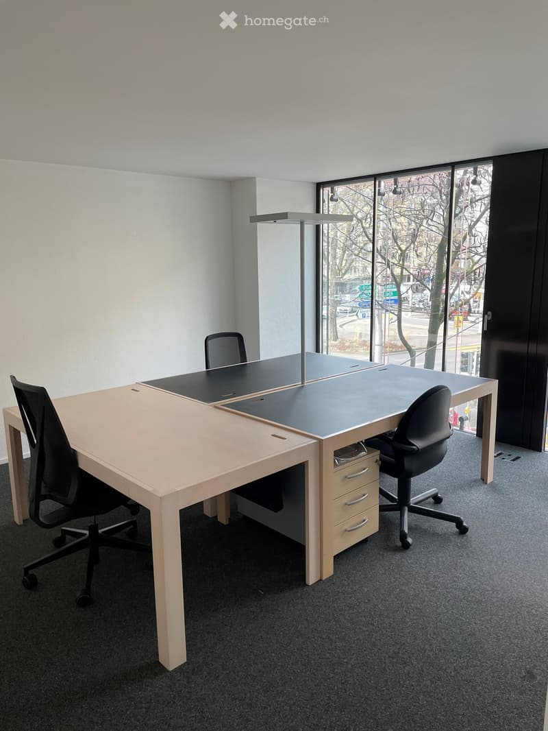 An empty meeting room with glass walls and large windows, two large wooden desks, four chairs, a lamp above the table, and a wooden drawer.