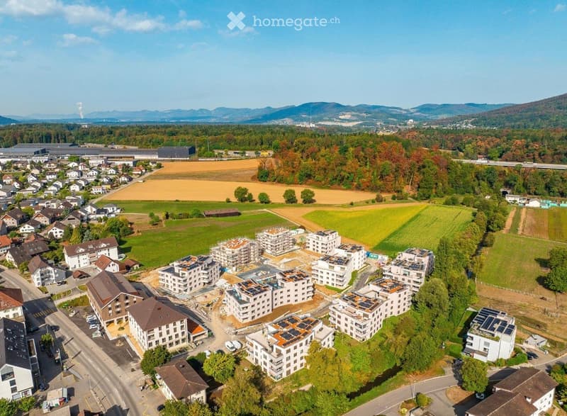 Aerial view of a residential area with multiple apartment buildings, some under construction, surrounded by green fields, trees, and distant mountains.