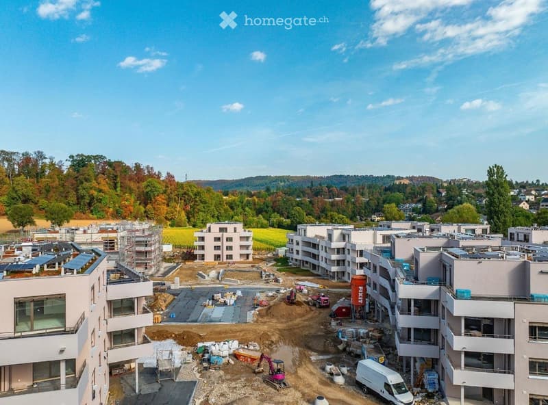 Aerial view of a residential construction site with multiple apartment buildings in various stages of completion, surrounded by greenery and a clear blue sky.
