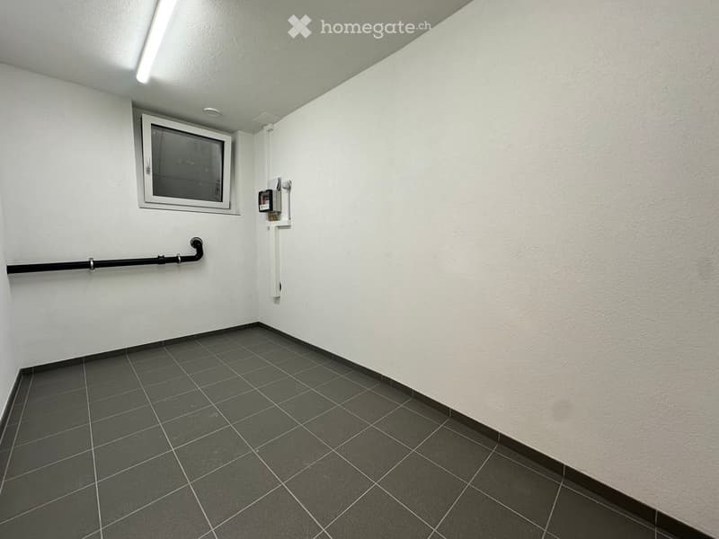 Empty room with white walls, grey tiled floor, and a small window with a metal frame. There is a black pipe and a white electrical panel on the wall.