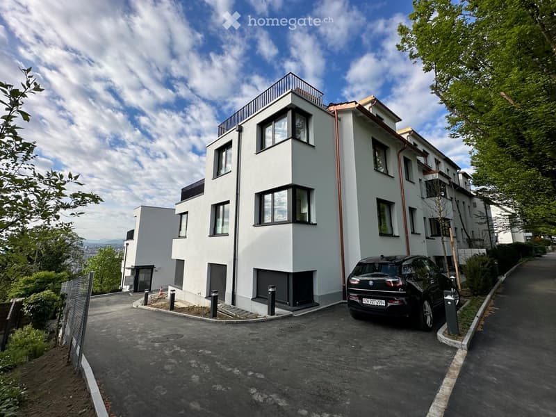 Modern multi-story residential building with white facade, large windows, and a rooftop terrace. A black car is parked in the driveway.