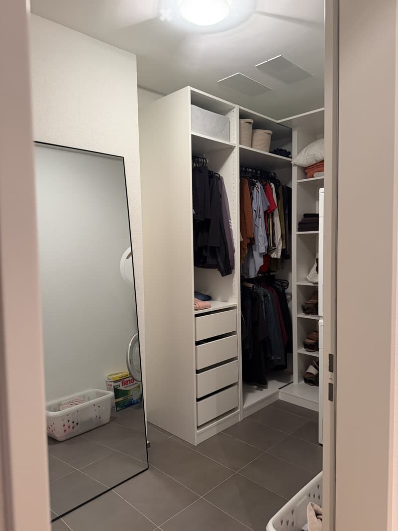 Well-organized walk-in closet with white built-in wardrobe, multiple shelves, hanging space for clothes, and drawers. Includes a full-length mirror and laundry baskets.