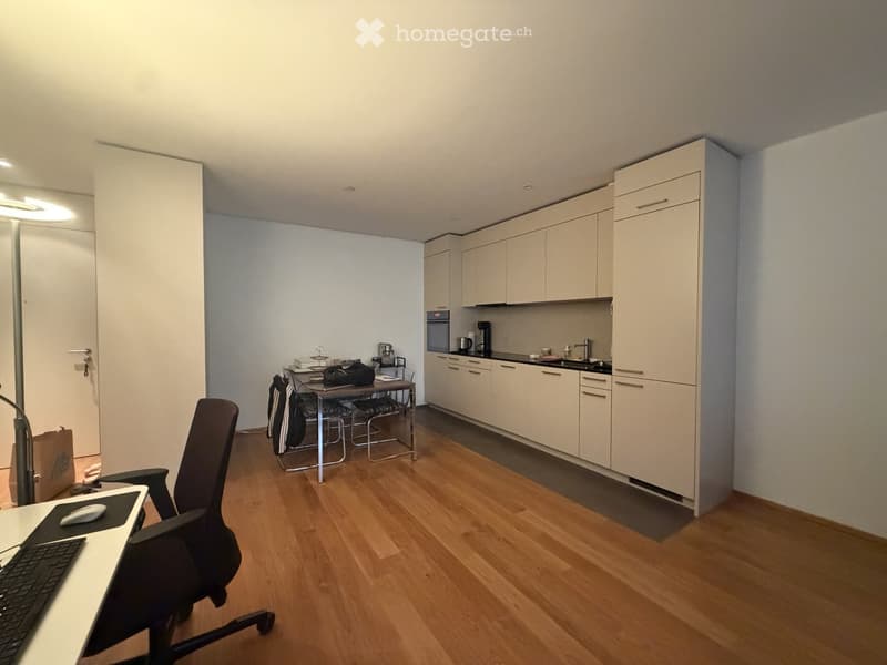 A modern, well-lit room with a wooden floor, a desk with a laptop and chair, a small kitchen area with cabinets, sink, and appliances, and a dining table with chairs.