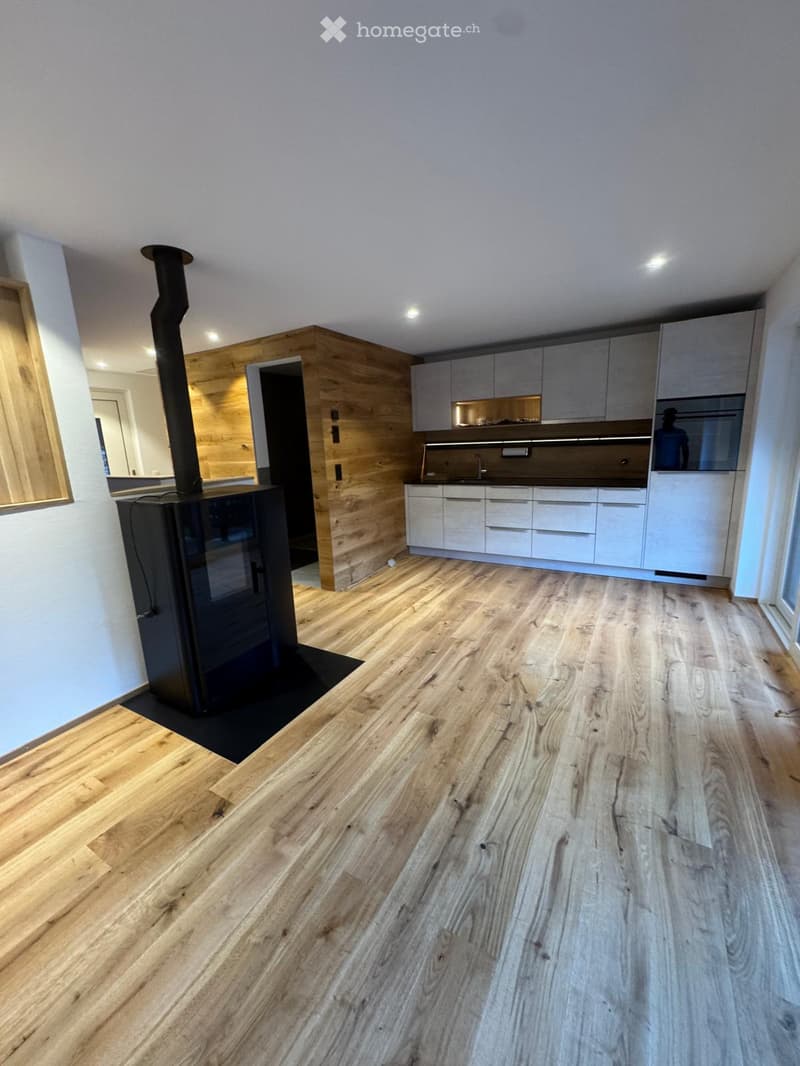 Empty room with light wooden flooring, white walls, and a modern kitchen area with white cabinets and stainless steel appliances visible in the background. A wood-burning stove is present on the left side.