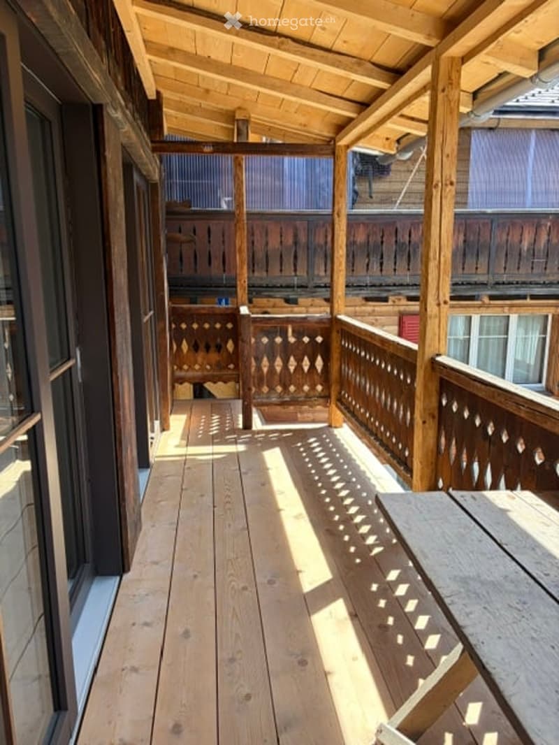 Wooden balcony with slatted floor, decorative wooden railings, and a wooden table on the right side.