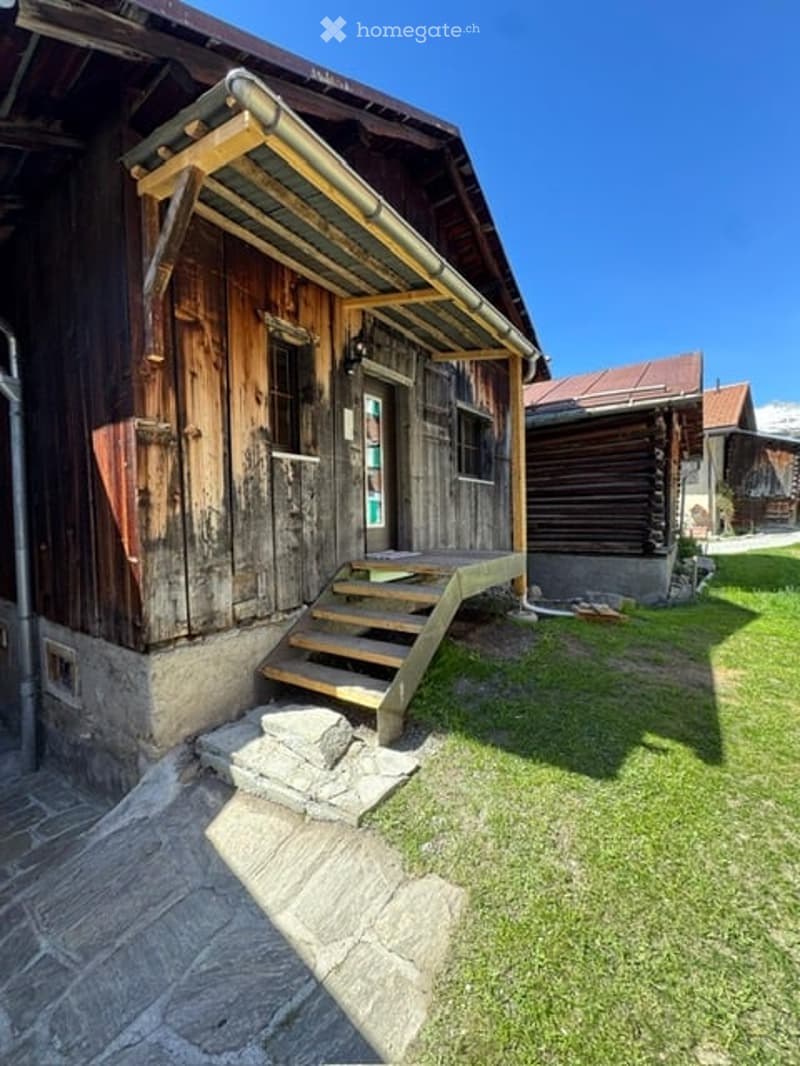 A rustic wooden house with a stone base, wooden steps leading to a covered entrance, and a grassy yard.