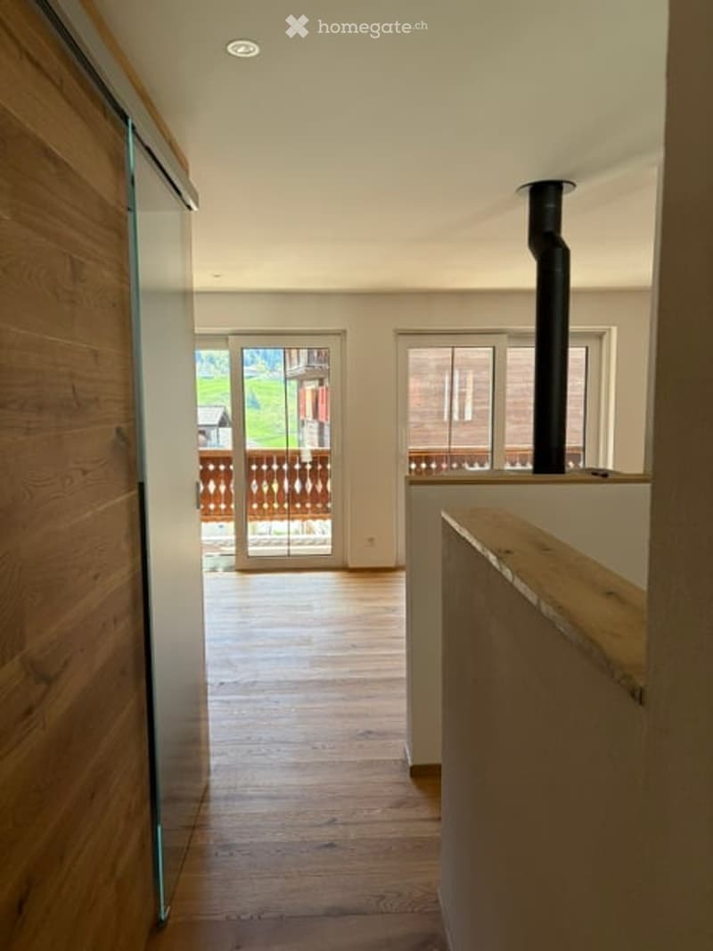 Bright hallway with wooden flooring, white walls, and large windows leading to a balcony with wooden railings.