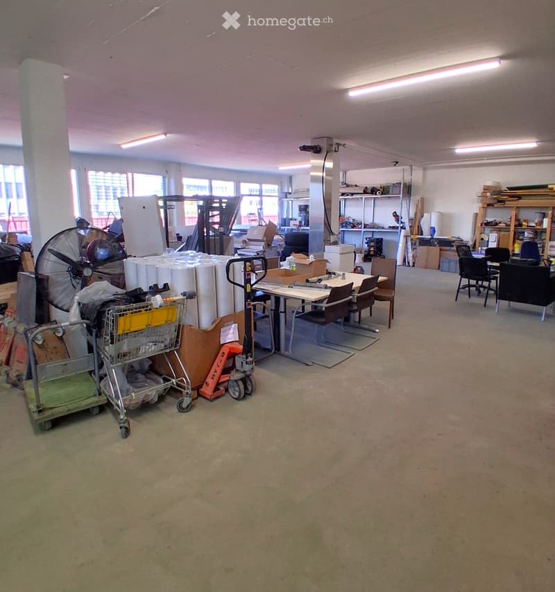 An empty warehouse with various items and equipment scattered around, including a fan, a trolley, a desk, and chairs.