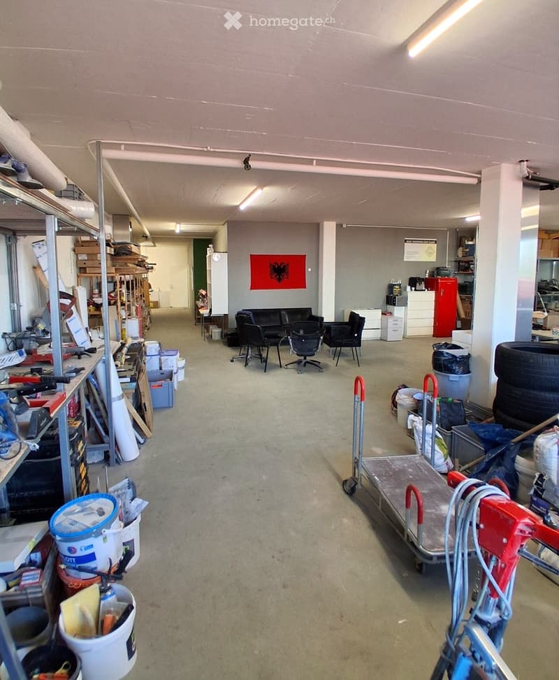 An empty room with a concrete floor, metal shelves, and various items scattered around. There are buckets, storage boxes, and tools on the shelves. There are also trolleys, tires, and other items on the floor.