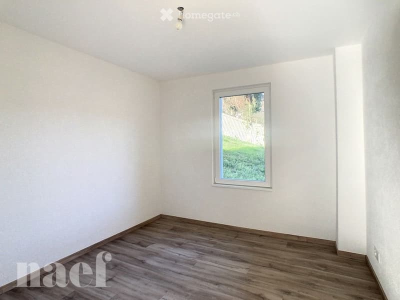 Empty room with wooden flooring, white walls, and a single window showing a view of a green outdoor area.