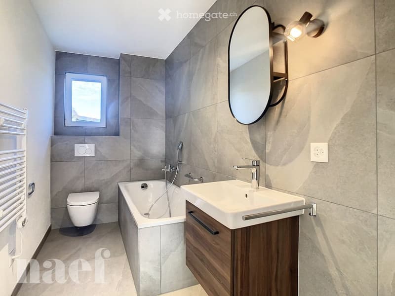 Modern bathroom with grey tiled walls, a bathtub, a sink with a wooden cabinet, a toilet, a large oval mirror, and a radiator.