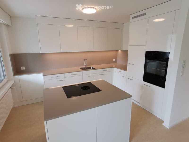 Modern, open-concept kitchen with white cabinets, beige countertops, built-in appliances including a dishwasher, and a central island with a cooktop.