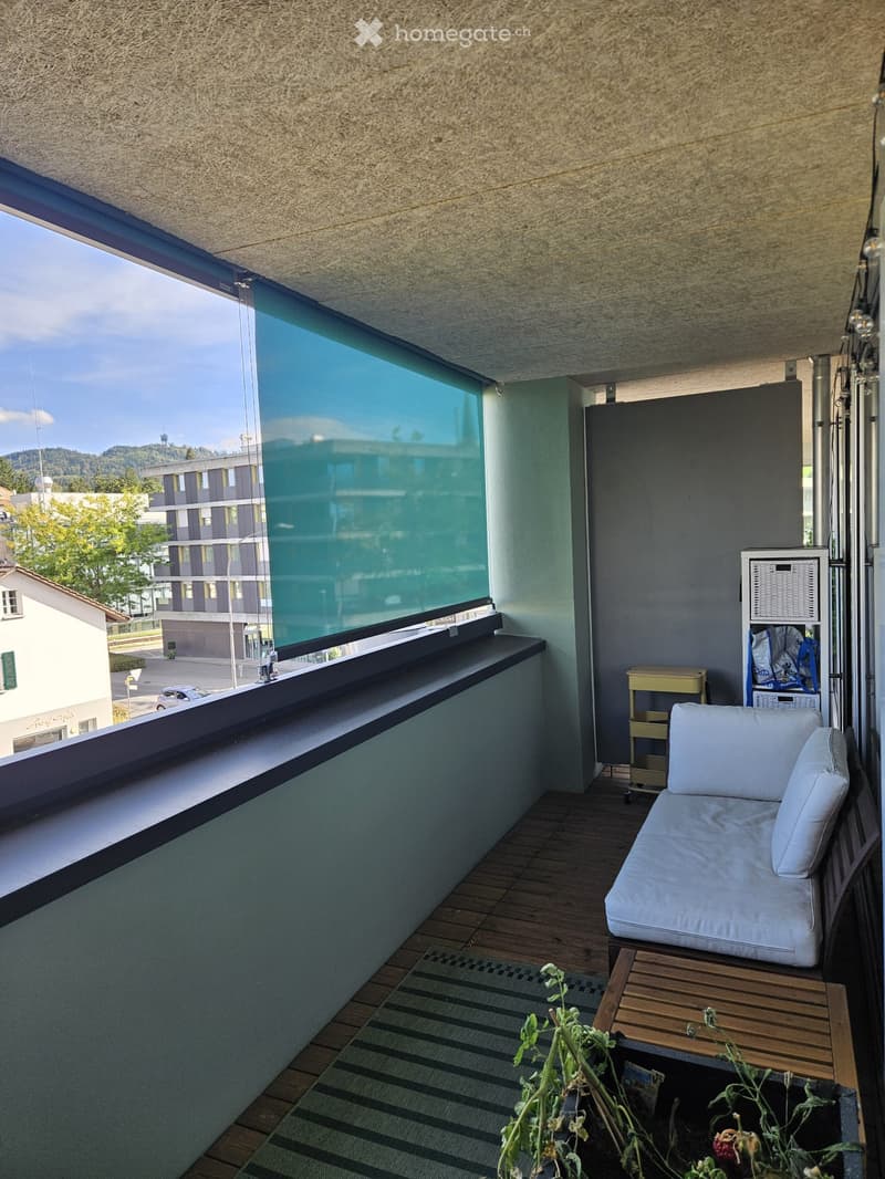 Balcony with wooden flooring, a white sofa, a small wooden table, and a storage unit. Large glass window offers a view of the city and surrounding landscape.