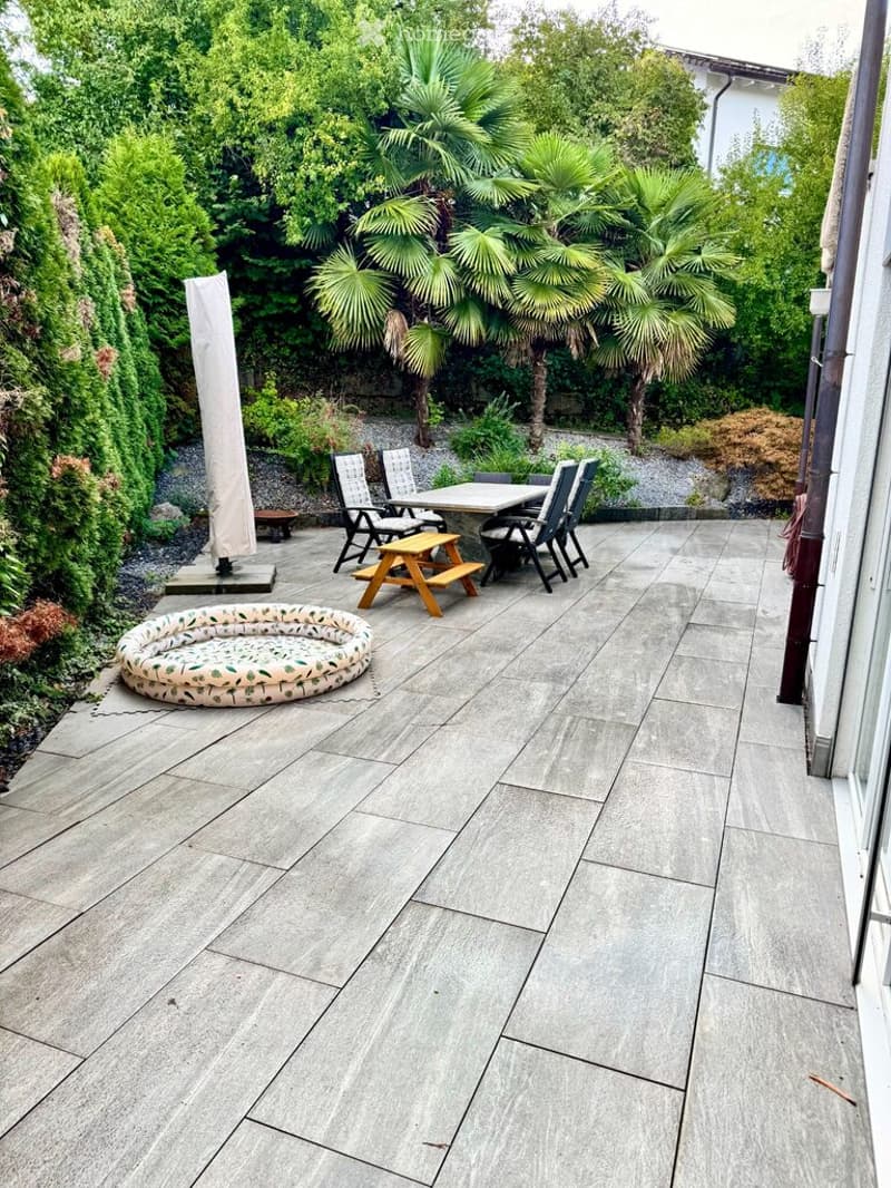 A modern patio with grey stone tiles, featuring a round inflatable pool, a wooden picnic table with chairs, and a covered umbrella stand. The area is surrounded by lush greenery and palm trees.