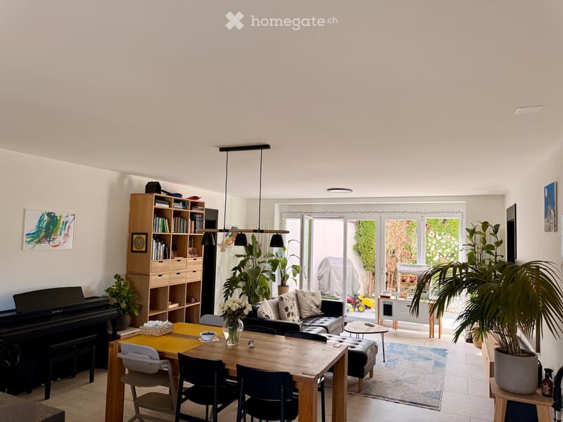 Bright living and dining area with a sofa, dining table, chairs, piano, bookshelf, and large windows leading to a patio.