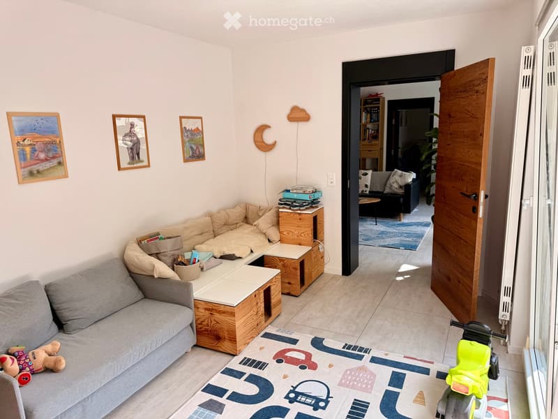 Living room with a grey sofa, wooden coffee table, bookshelf, and a small table with books and a basket. A toy scooter and a stuffed toy are also present.