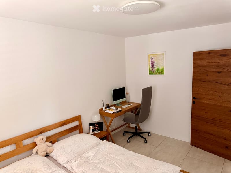 A small, well-lit bedroom with a wooden bed frame, a desk with a computer, a chair, and a nightstand with a lamp and decorative items.