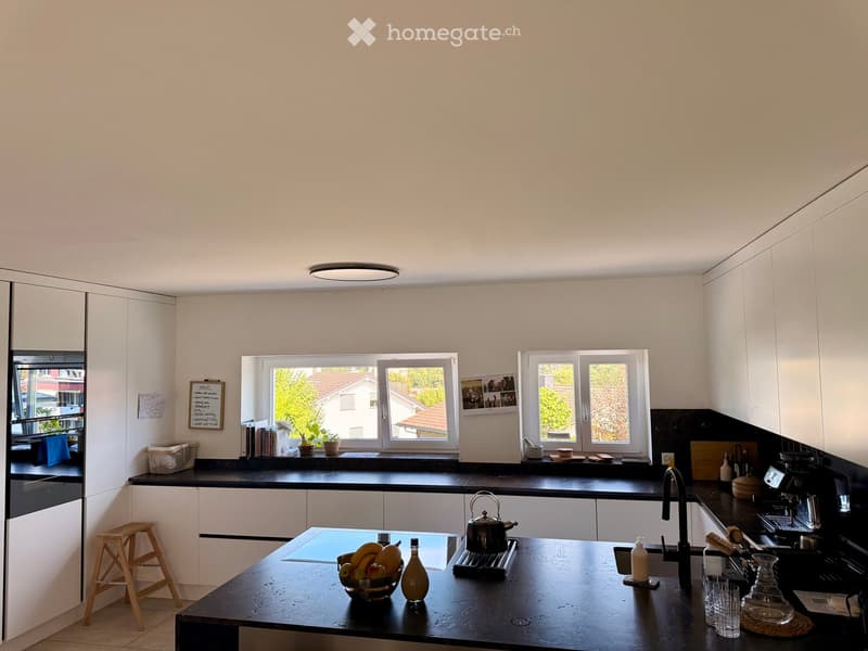 Modern kitchen with white cabinets, black countertops, and various appliances. Features include a central island with a bowl of fruit, a teapot, and a coffee maker, along with a small wooden stool and a window view of the outdoors.
