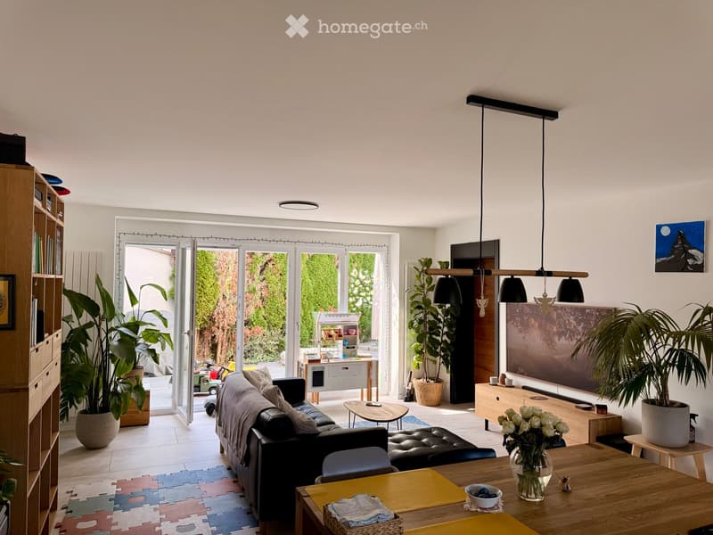 Bright living room with large windows and sliding doors leading to a garden. Features a black leather sofa, ottoman, coffee table, dining table with chairs, potted plants, and a shelf with books and decorative items.