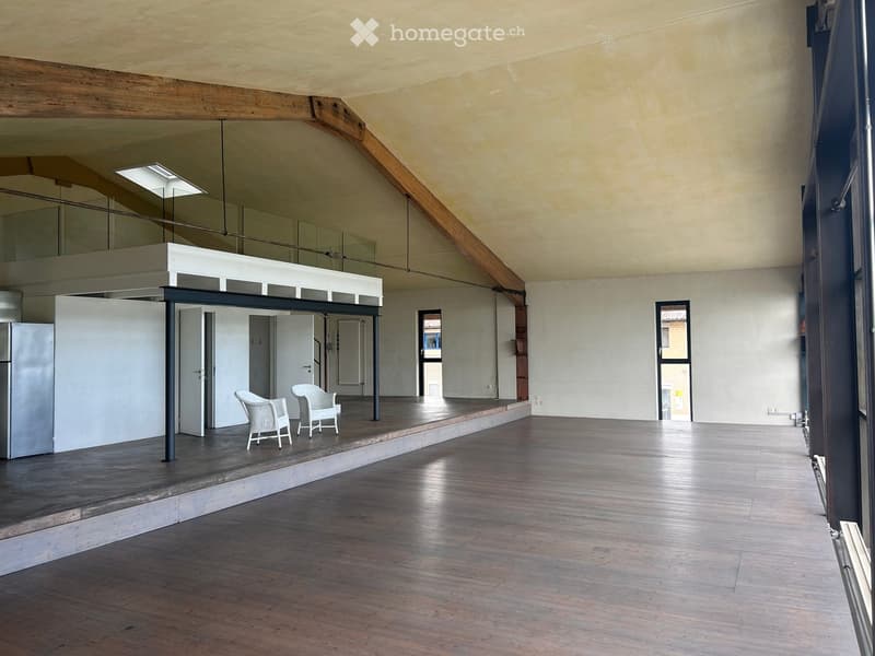 Open, spacious room with wooden flooring, exposed wooden beams, and a raised platform area with two white chairs and a refrigerator. The room features large windows and a modern, minimalist design.