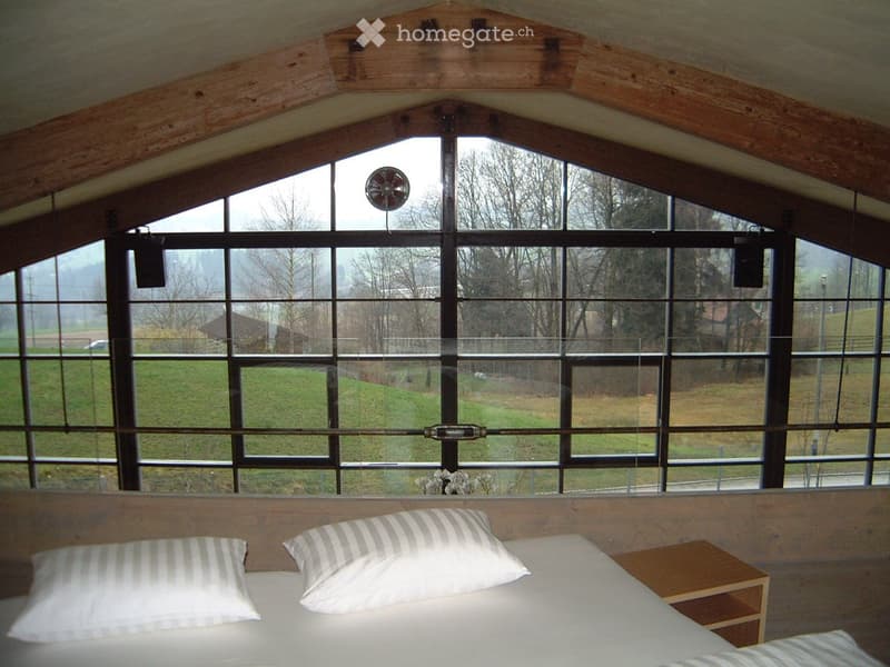 Bright bedroom with large windows, wooden ceiling beams, a bed with white bedding and striped pillows, and a small wooden nightstand.