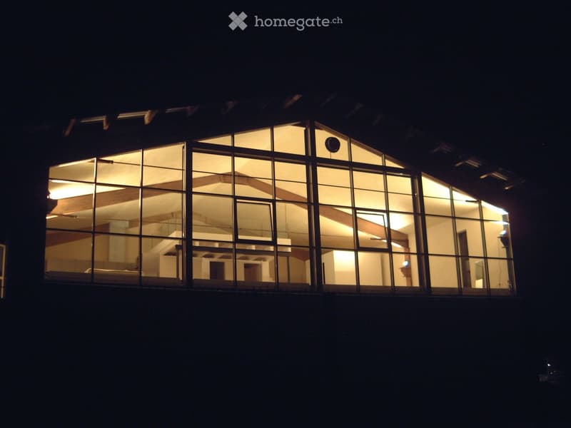 Nighttime view of a modern building with large glass windows, interior lights visible, and a curved architectural design.