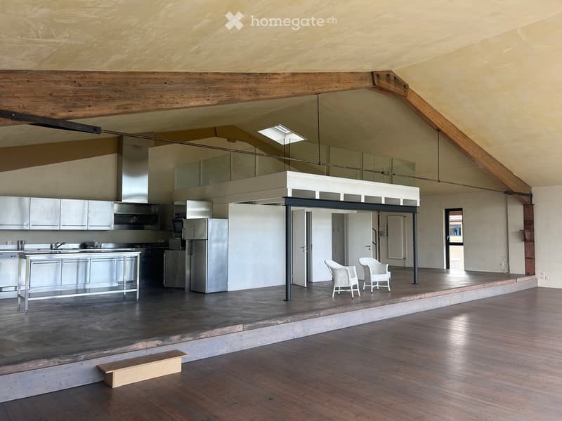 Open-plan space with a modern kitchen area featuring stainless steel appliances and a kitchen island, and a seating area with two white chairs and a small wooden stool.