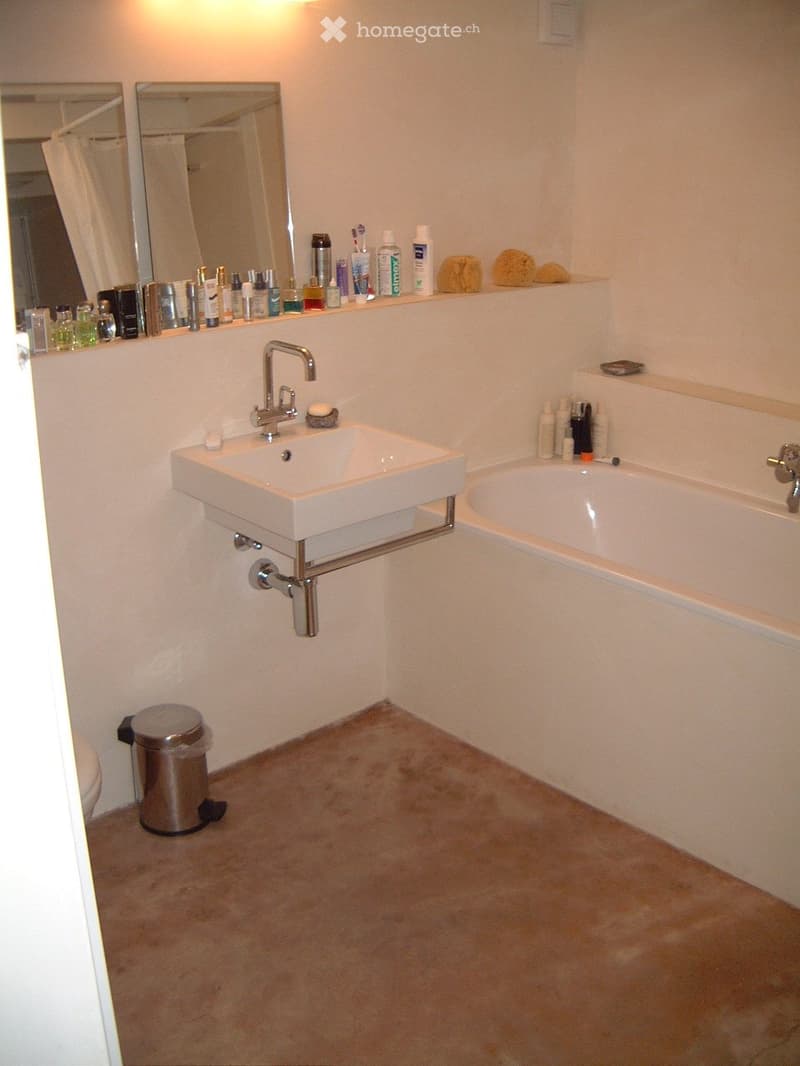 Bathroom with a white sink, faucet, bathtub, mirror, and various toiletries on the counter. A trash can is placed near the toilet.