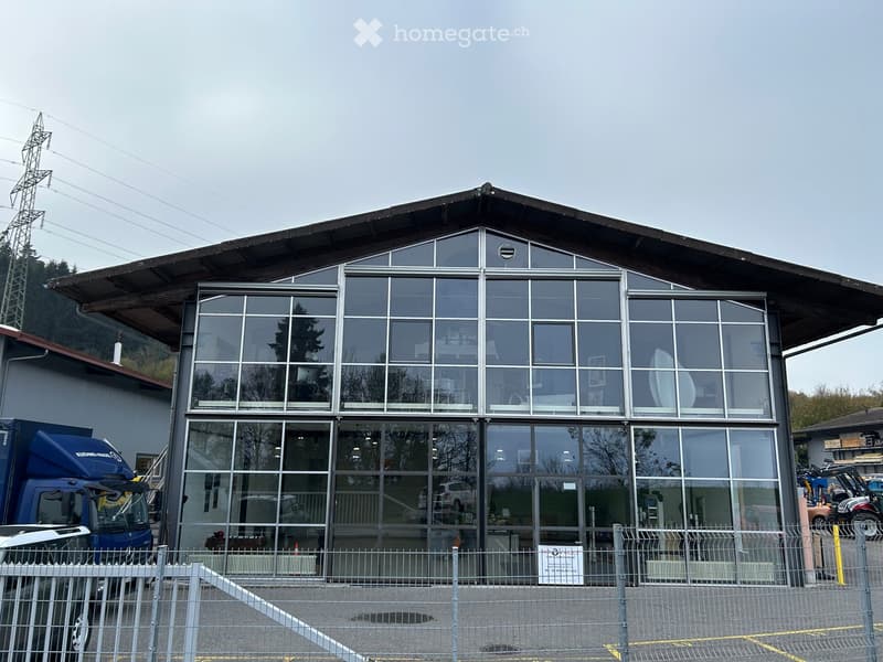 Modern building with large glass windows and a sloped roof, surrounded by a metal fence and parked vehicles.