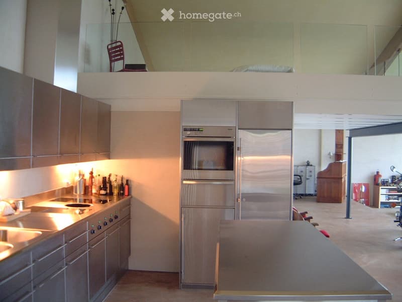 Modern kitchen with stainless steel appliances, wooden cabinets, and a sleek countertop. A small seating area with a chair is visible in the background.