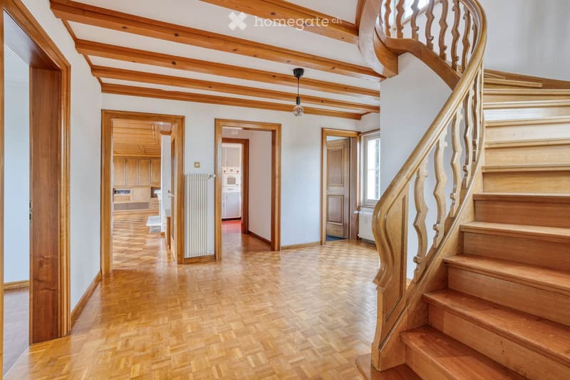 Spacious hallway with wooden flooring, exposed wooden beams on the ceiling, and a wooden staircase with a carved railing leading upwards. Several doors open to adjacent rooms, including a kitchen area visible in the background.